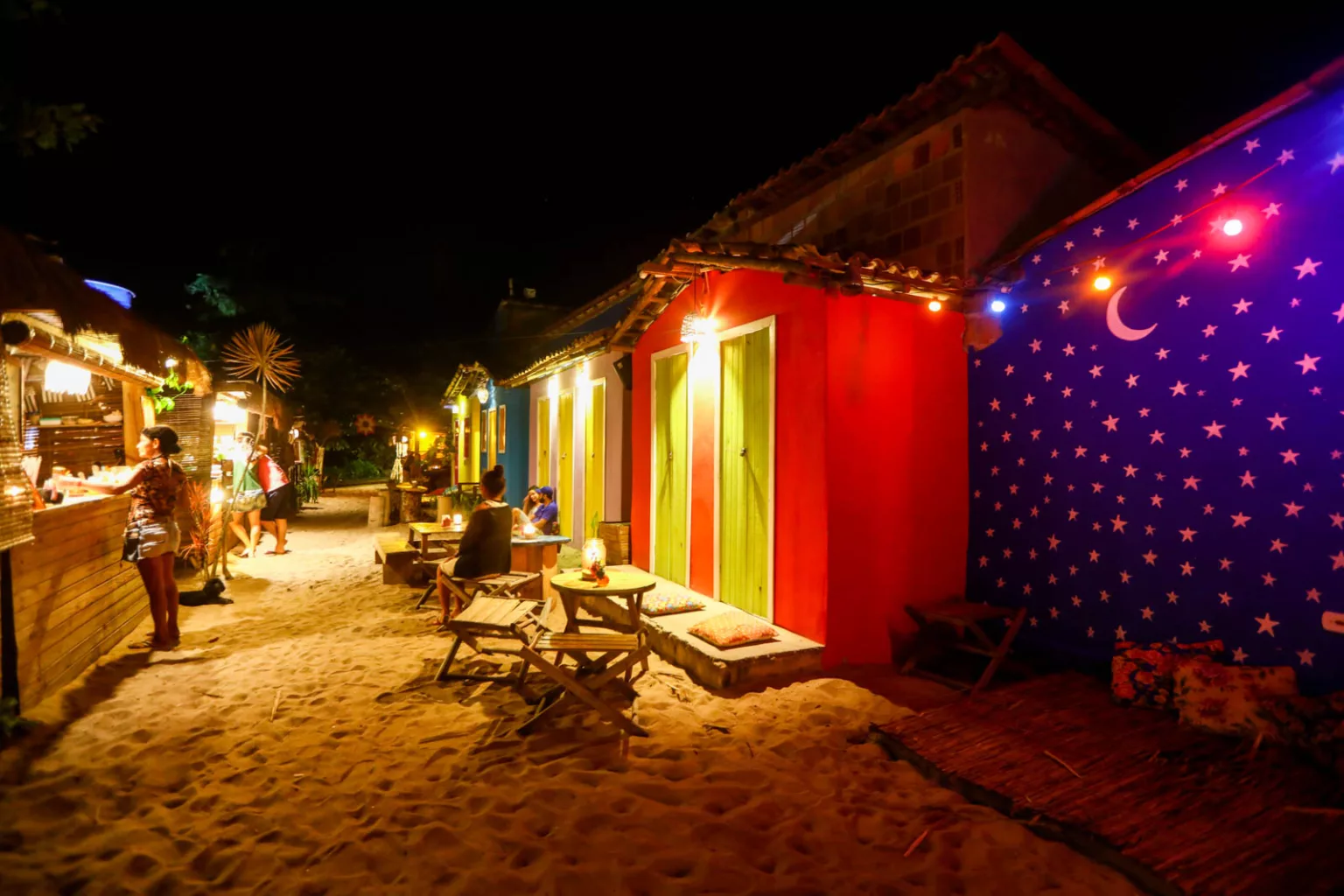 Ruas de areia de Caraíva iluminadas à noite, com bares e casinhas coloridas